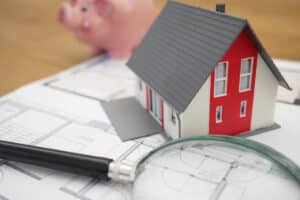 Modèle de maison rouge sur des plans architecturaux avec une loupe et une tirelire, représentant des économies sur l'assurance emprunteur.