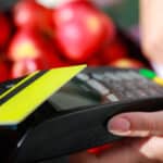 Main insérant une carte jaune dans un terminal de paiement avec des pommes rouges en arrière-plan.